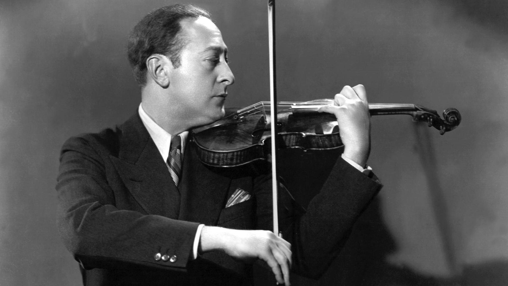 Schwarz-weiß-Fotografie, sie zeigt den Geiger Jascha Heifetz im Profil, er spielt Geige und führt den Bogen mit der rechten Hand. Er hat kurz dunkle Haare, trägt einen schwarzen Anzug mit weißem Hemd und Schlips
