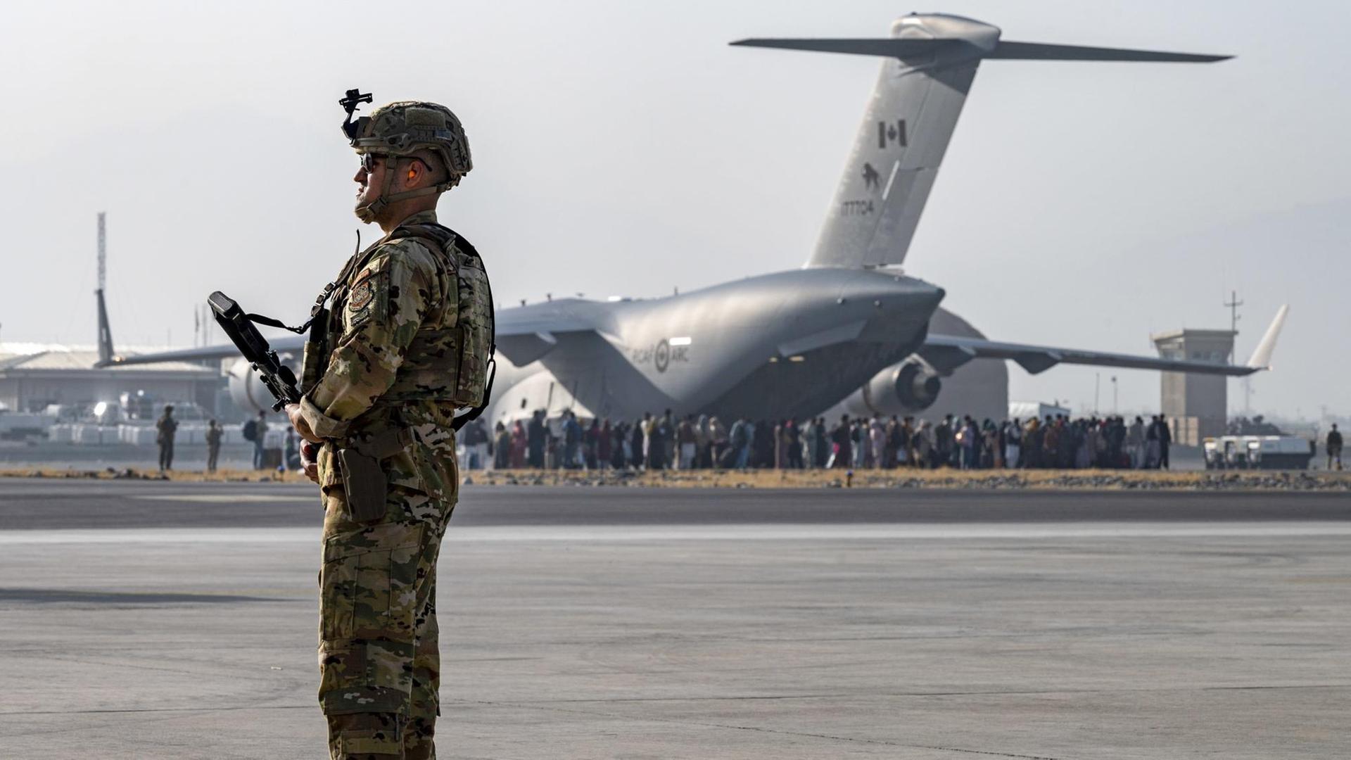 Ein bewaffneter US-Soldat steht am Flughafen Kabul, im Hintergrund ein Flugzeug, das Menschen aus Afghanistan ausfliegen soll.