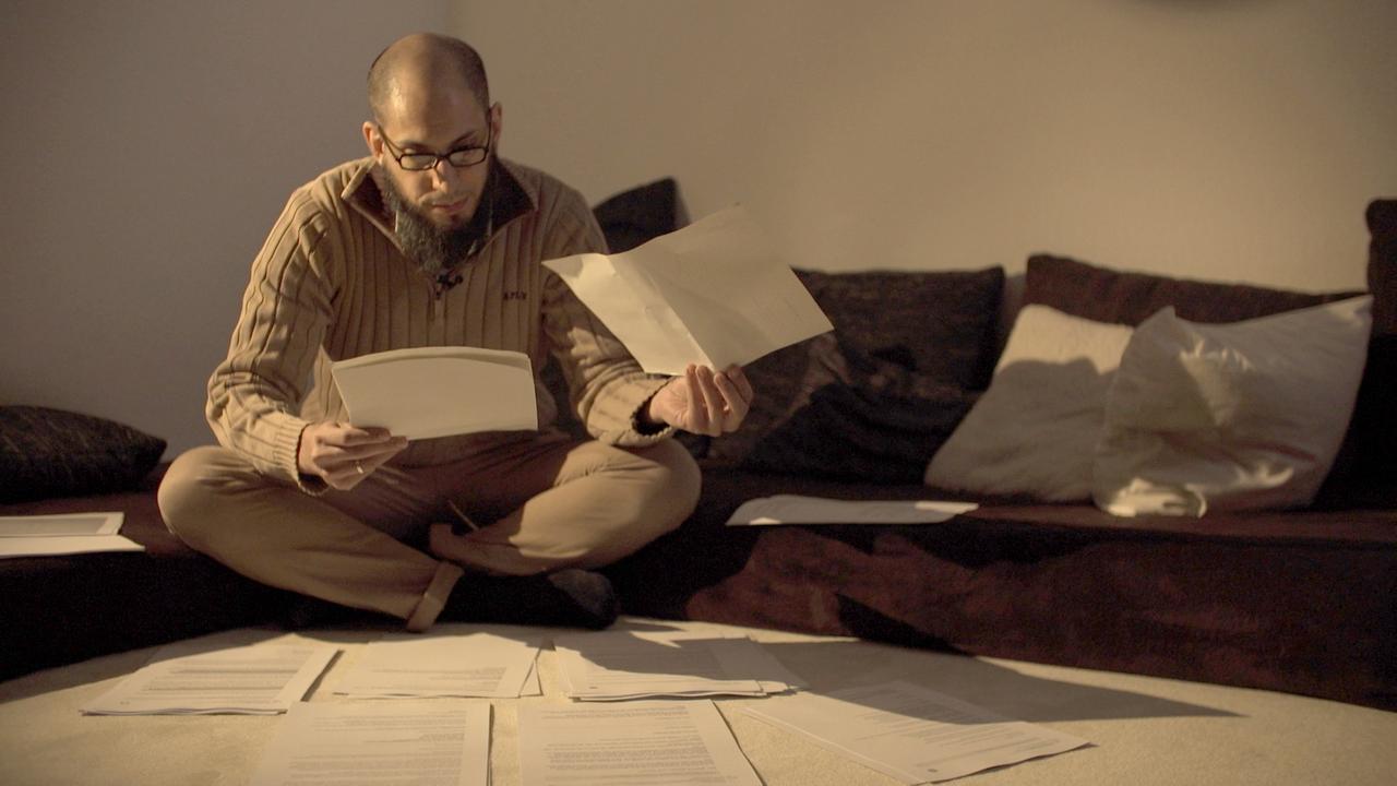 Der Protagonist Danisch sitzt auf dem Boden vor verschiedenen Dokumenten, einige hat er in der Hand und liest sie.