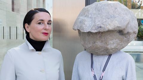 Das Bild zeigt die Künstlerin Natascha Süder Happelmann mit einer Steinskulptur auf dem Kopf, die ihr Gesicht verbirgt, und ihre Sprecherin Helene Duldung.