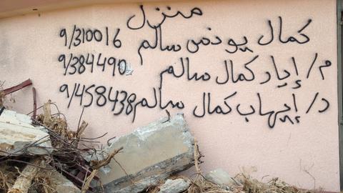 Arabische Schriftzeichen und Zahlen stehen auf auf einer Hauswand, davor liegen Steine und Trümmer