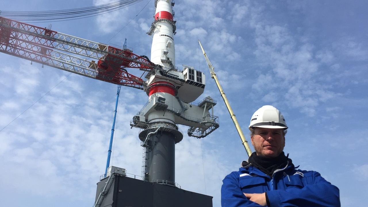 Projektleiter Keno Dirks mit einem Schutzhelm vor dem neuen Super-Kran im Rostocker Hafen.