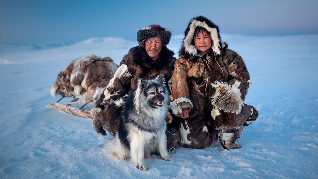 Zwei Tschuktschen posieren mit ihrem Hund im Schnee, im Hintergrund ein mit Fellen bedeckter Schlitten.