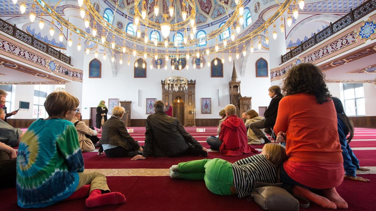 Die Besucher sind von hinten zu sehen, wie sie auf dem roten Teppich des Gebetsraums sitzen. Über ihnen leuchtet ein riesiges hängendes Lampenemseble.