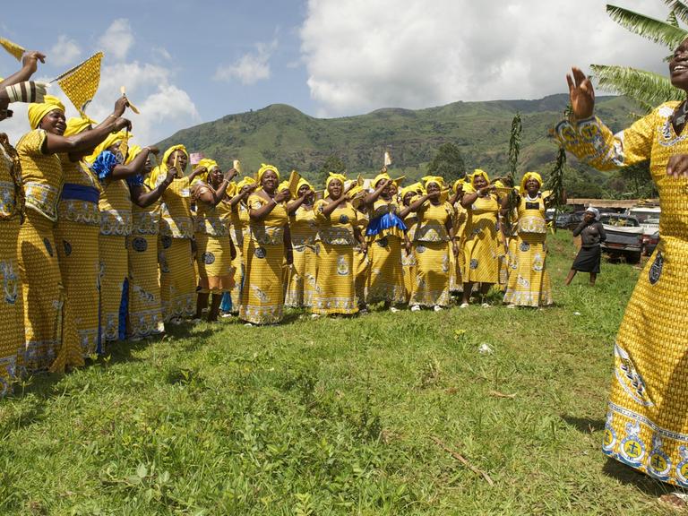 Frauen nehme am "Christian Women's Fellowship" in Kamerun teil. Sie tragen gelbe Gewänder, die Leiterin der Gruppe steht vor ihnen und gibt Handzeichen.
