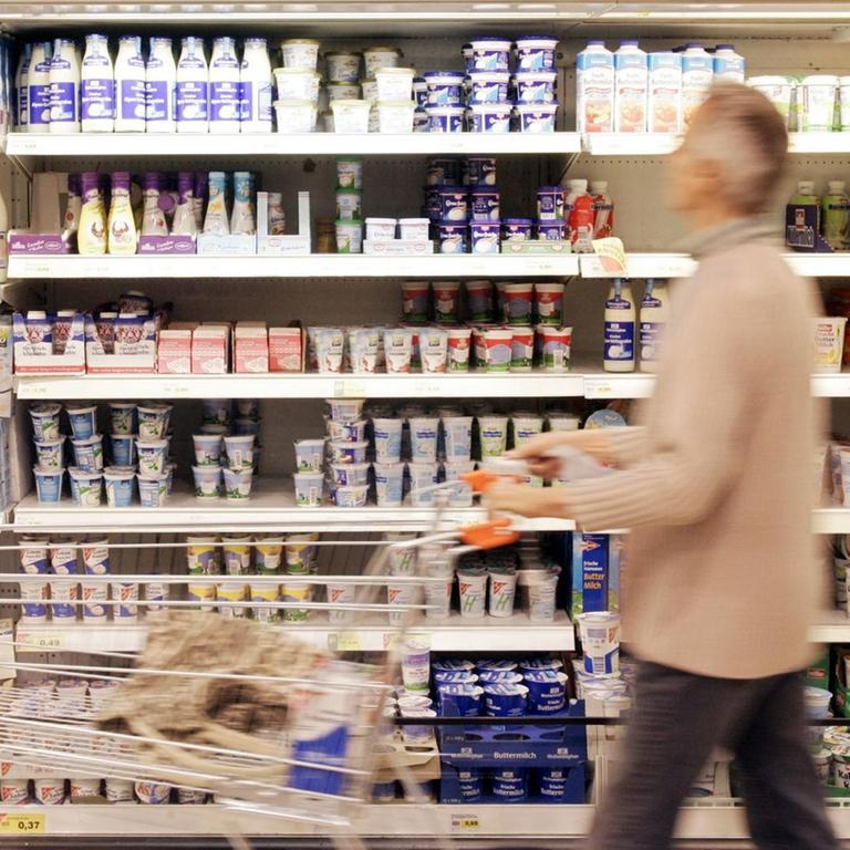 Ein Supermarktregal mit Plastikverpackungen. Davor geht ein grauhaariger Mann mit einem Einkaufswagen.