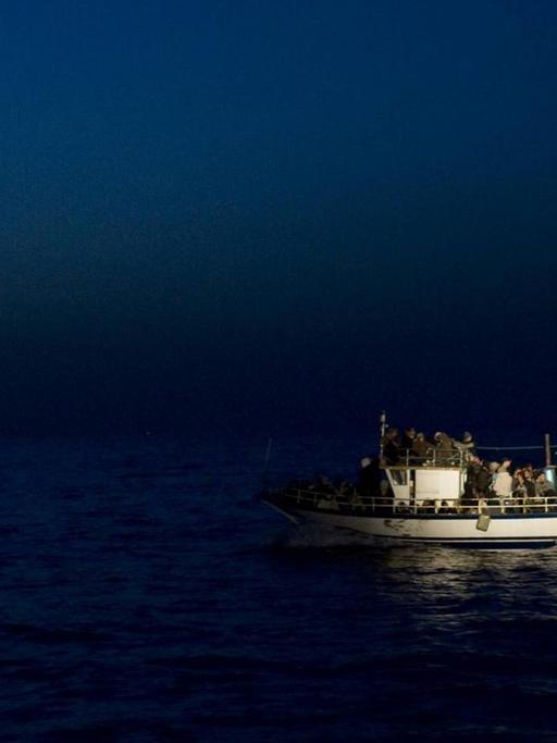 Ein kleines Boot mit vielen Menschen liegt auf dem Wasser. Es ist Nacht, das Boot wird angestrahlt.