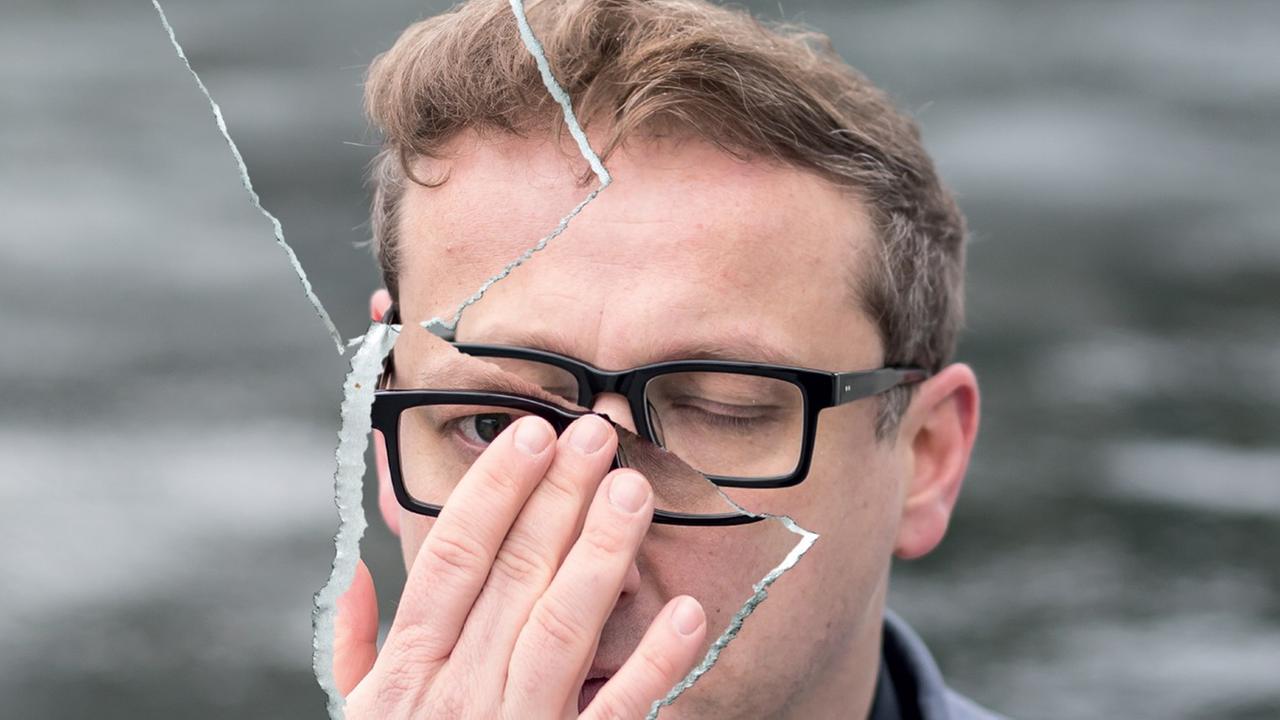 Ein Mann mit schwarzer Brille schaut durch eine zerbrochene Scheibe. Er hält die rechte Hand vor sein Gesicht.