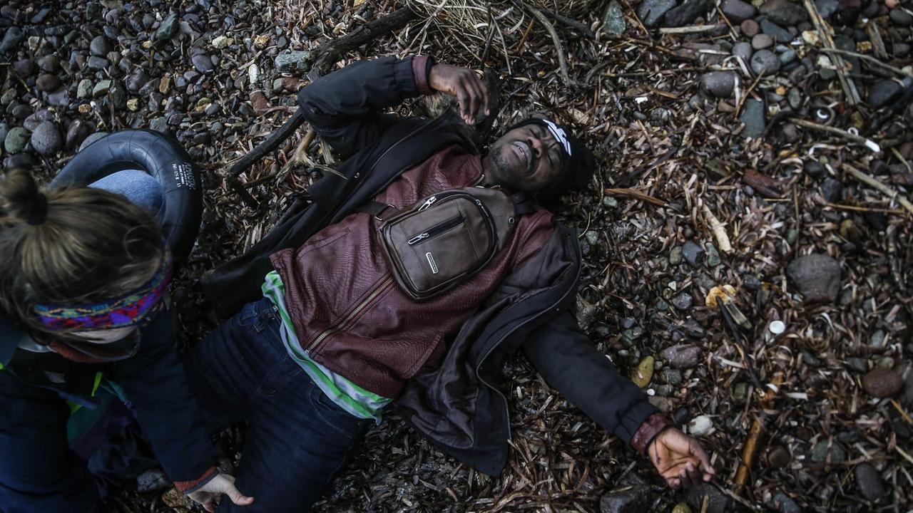 Ein Mann liegt erschöpft am Steinstrand von Lesbos während eine Frau sein linkes Bein abtastet.