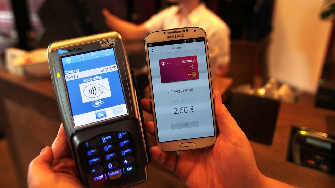Eine Frau zeigt ein Smartphone mit der App MyWallet neben einem Empfangsgerät an der Kasse eines Restaurants.