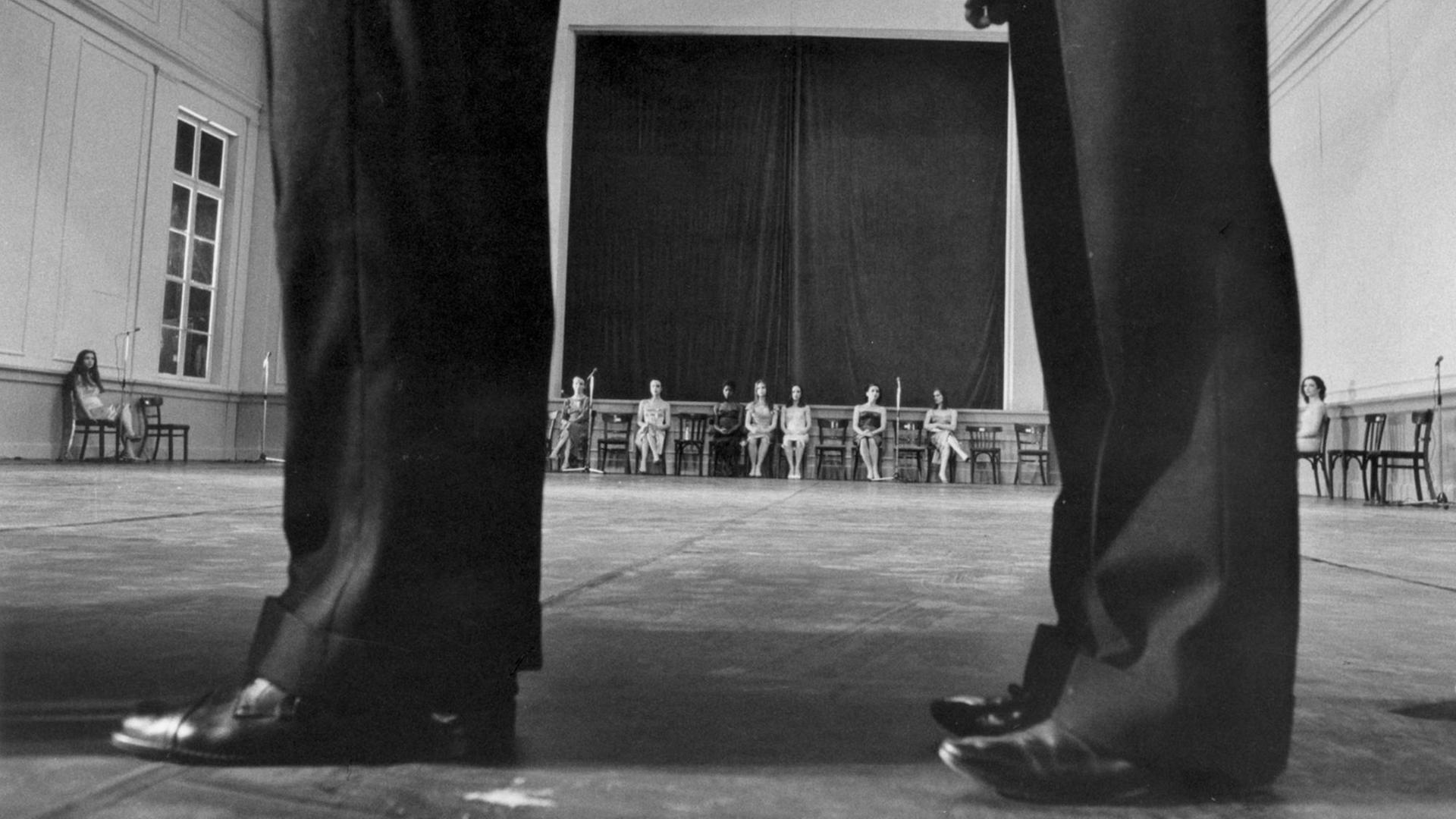 Im Vordergrund zwei Paar Beine in schwarzen Hosen. Blick durch die Beine nach hinten, wo mehrere Frauen auf Stühlen sitzen. Schwarzweißfoto.