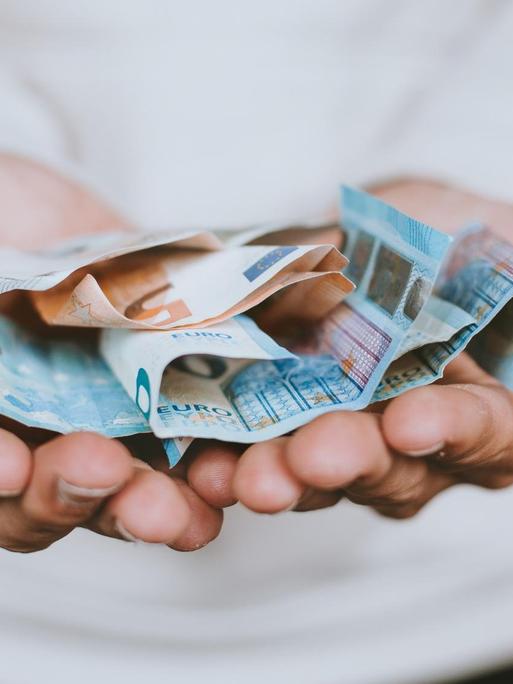 Eine Person im weißen Pullover hält die Hände zu einer nach oben geöffneten Schale. Darin liegen mehrere 20-Euro-Scheine und ein 50-Euro-Schein.