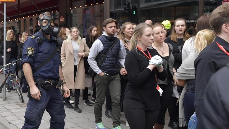 Das Bild zeigt einen Polizisten mit Gasmaske und eine größere Menschenmenge in Stockholm