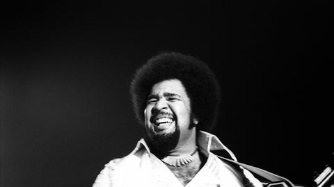 Das s/w-Bild zeigt einen Mann mit Vollbart und Afro, der ein Umhängekeyboard hält. Er blickt lachend in die Ferne.