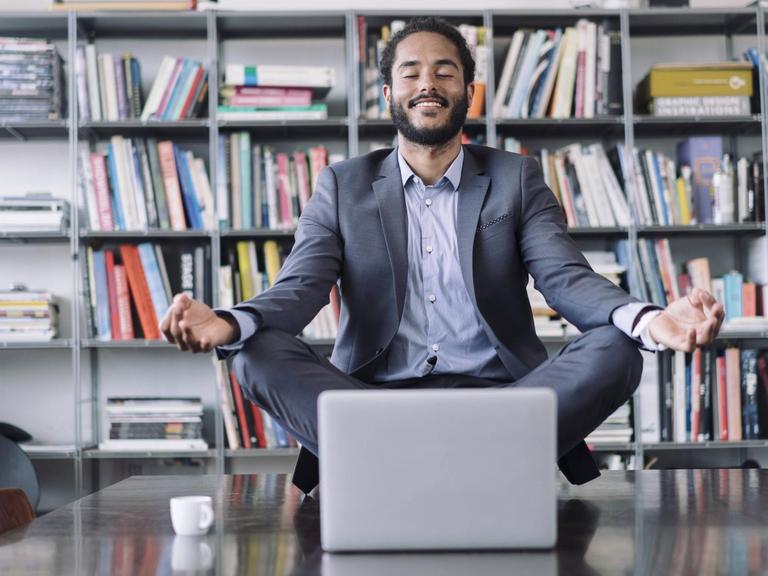 Ein junger Geschäftsmann sitzt in Meditationshaltung auf seinem Arbeitstisch hinter seinem Laptop