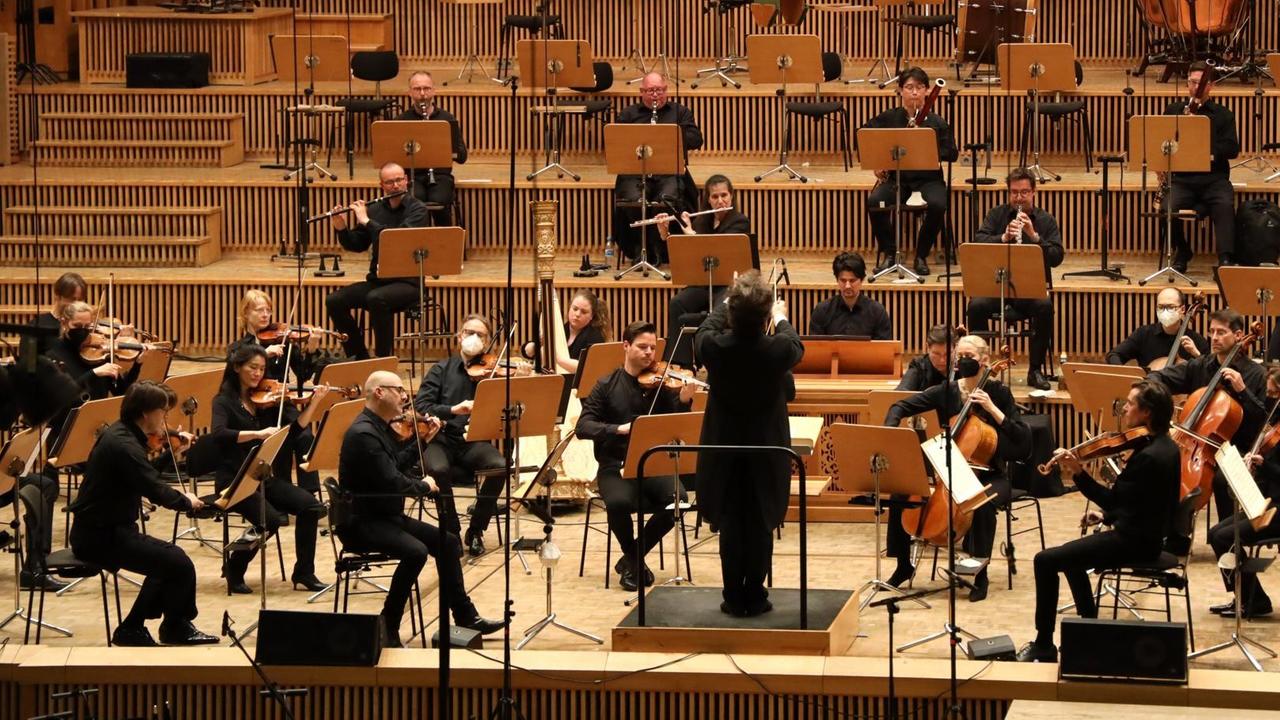 Ein Dirigent steht vor einem Orchester, das relativ weit auseinander und auf Stufen sitzt, die sich nach oben hin öffnen.