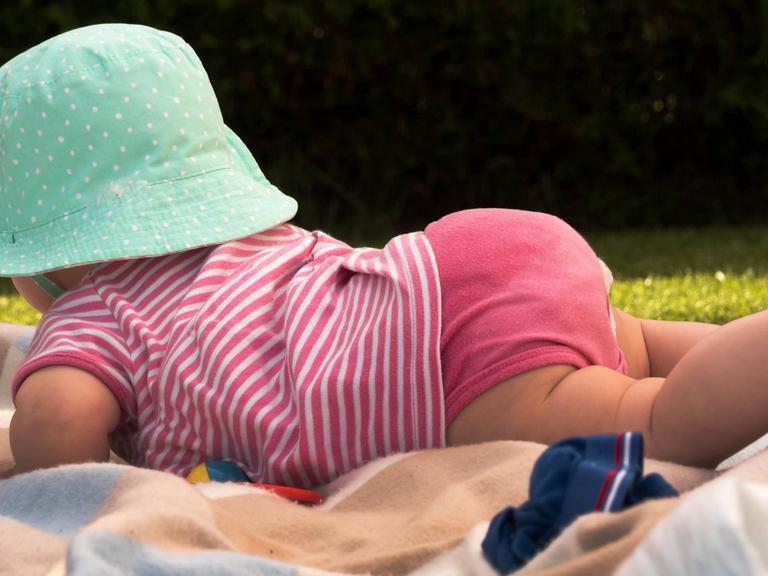 Ein Baby mit einem großen Sonnenhut liegt auf dem am Bauch auf einer Decke auf einer Wiese im Sommer.