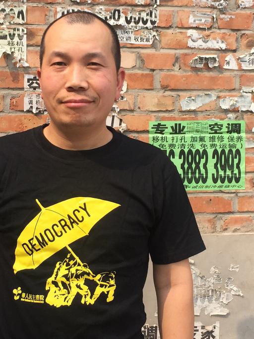 Der chinesische Künstler Lv Shang steht vor einer Backsteinwand und trägt ein schwarzes T-Shirt mit einem gelben Regenschirm, auf dem "Democracy" steht.