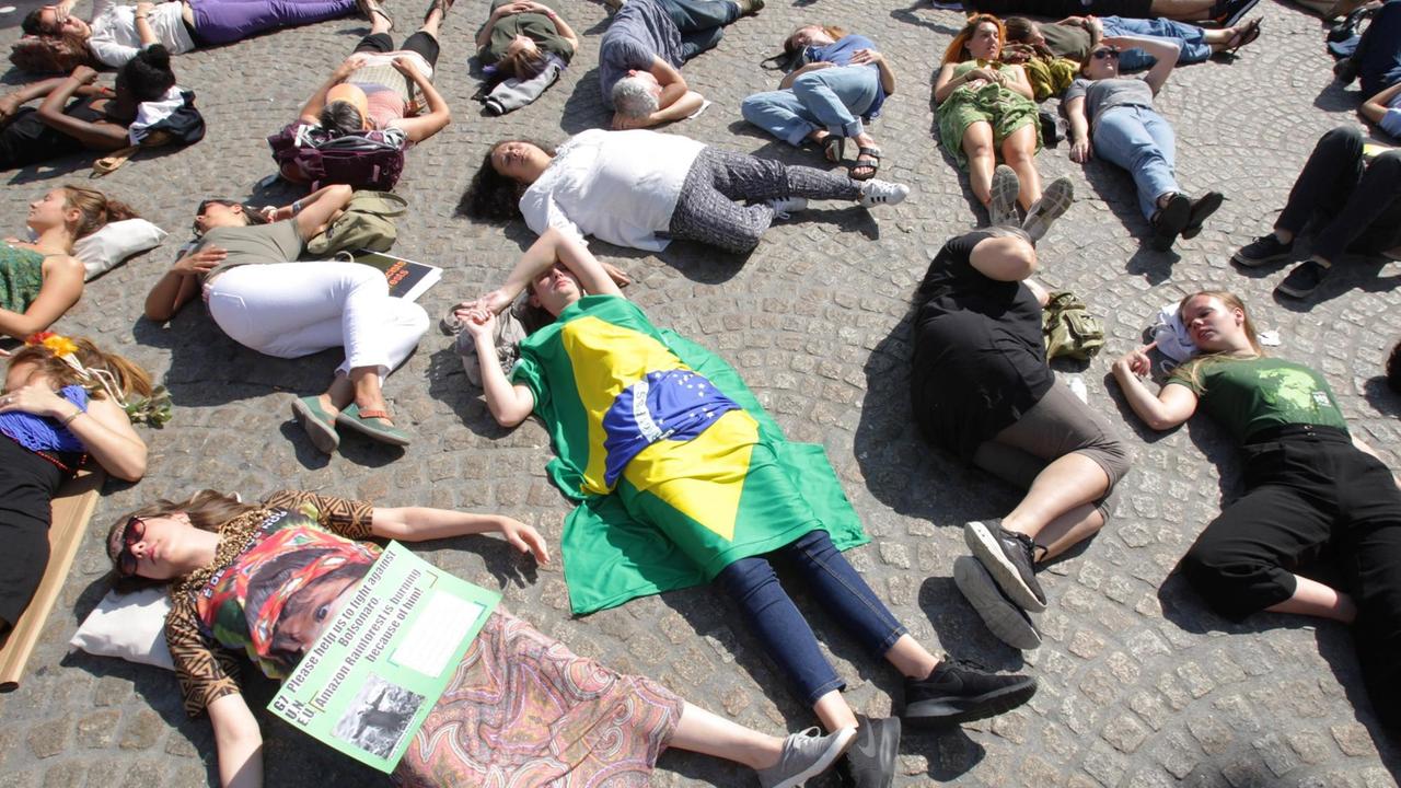 Menschen liegen während einer Demonstration gegen die Amazonas-Rodungen auf dem Boden, als ob sie gestorben seien. Eine Person ist in eine Brasilien-Flagge gehüllt, auf dem Schild einer anderen steht: Please help to fight against Bolsonaro. Amazon rainforest is burning because of him.