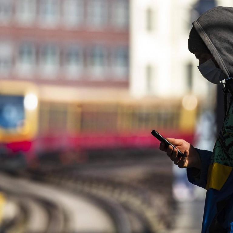 Ein Jugendlicher steht mit Mundschutzmaske auf einem S-Bahnsteig in Berlin und blickt auf sein Handy.