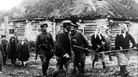 Frauen und alte Männer mit Mistgabeln sowie ein Partisan mit entsichertem Revolver gehen während des Zweiten Weltkriegs in einem russischen Dorf gegen einen Verräter vor, der Informationen an die Deutschen gegeben haben soll.