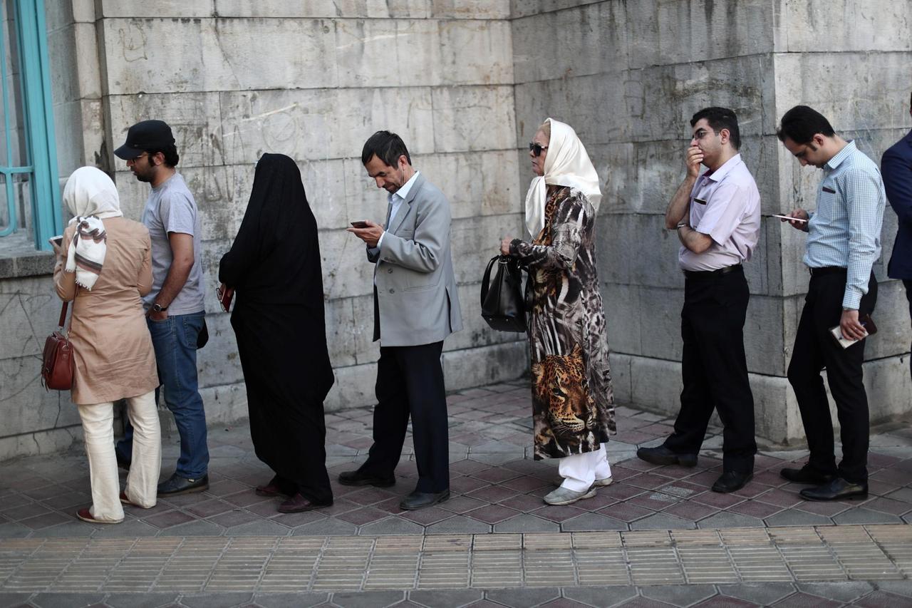 Mehrere Frauen und Männer stehen vor dem Gebäude in einer lockeren Warteschlange. Einige unterhalten sich, andere blicken auf ihre Smartphones.