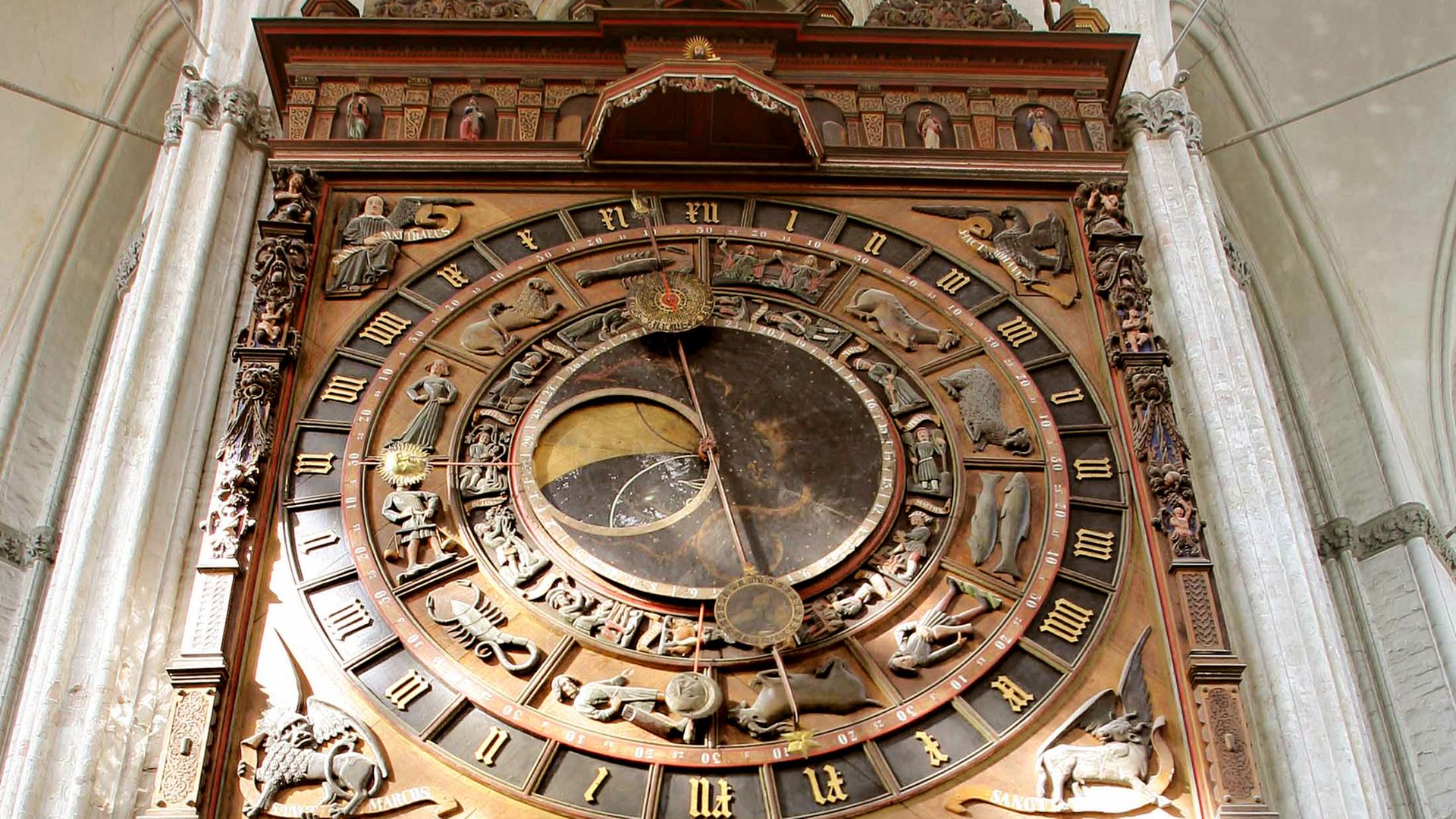 Eine alte astronomische Uhr von unten betrachtet in Großaufnahme. Zu sehen sind Holzschnitzereien, der Zeiger steht fast senkrecht.