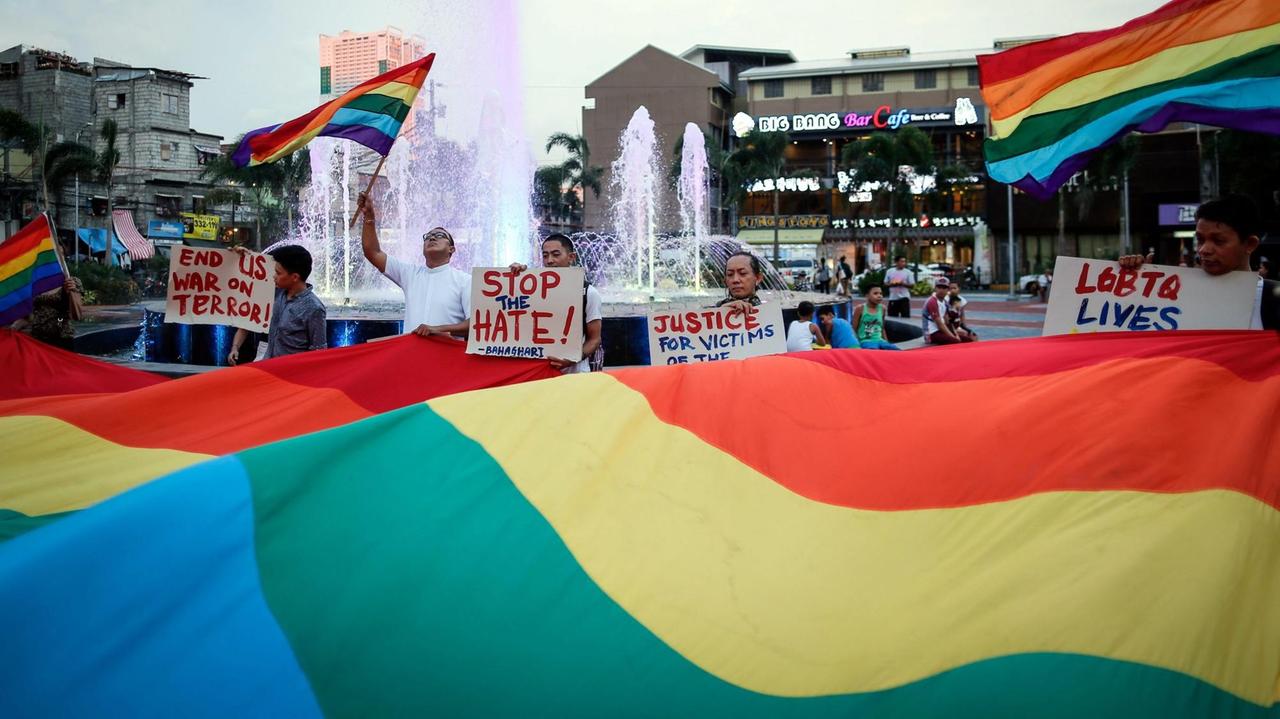 Demonstration von Angehörigen der LGBT-Community auf einem Platz mit Brunnen, es wehen Regenbogenflaggen, auf Plakaten steht "Stop the Hate"