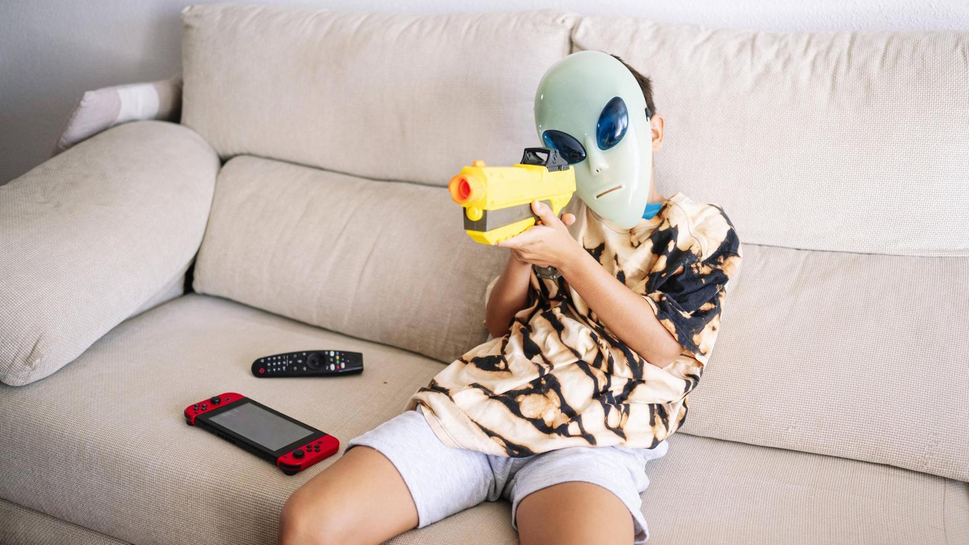 Ein Junge mit Alien-Maske und Plastikgewehr sitzt auf einem Sofa. file_source: imago / Westend 61