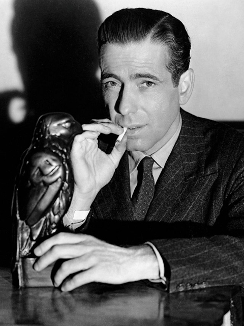 Humphrey Bogart als Sam Spade in "The Maltese Falcon" (1941). Schwarz-Weiß-Aufnahme von Humphrey Bogart, rauchend an einem Tisch sitzend.