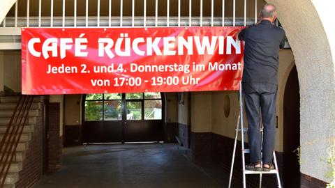 Stefan Friedrichowicz steht auf einer Leiter und hängt ein rotes Banner mit der Aufschrift "Cafe Rückenwind" auf.