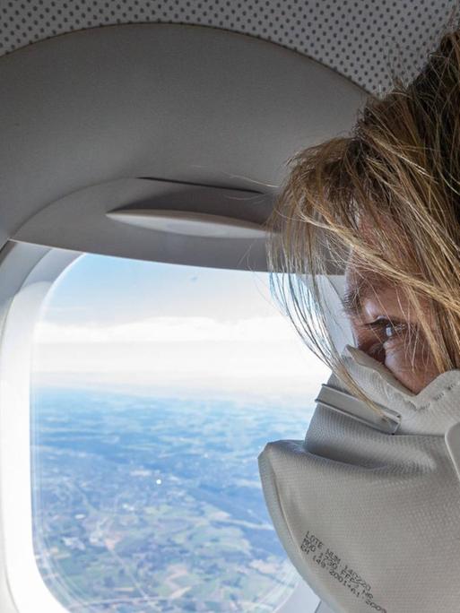 Eine Frau sitzt mit einer FFP2-Maske in einem Flugzeug am Fenster.