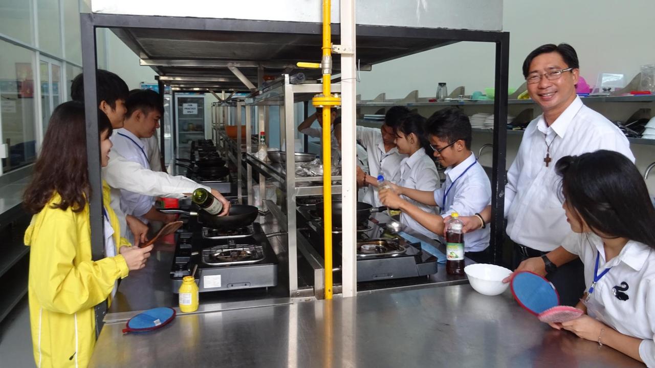 Ordensbruder Joseph Phan Trung Hoan gibt benachteiligten Teenagern Kochkurse in einer großen Küche. Jeder hat eine Pfanne in der Hand.