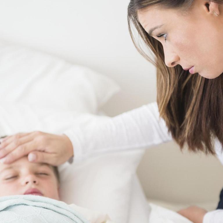 Ein kleiner Junge liegt schlafend im Bett, seine Mutter befühlt seine Stirn