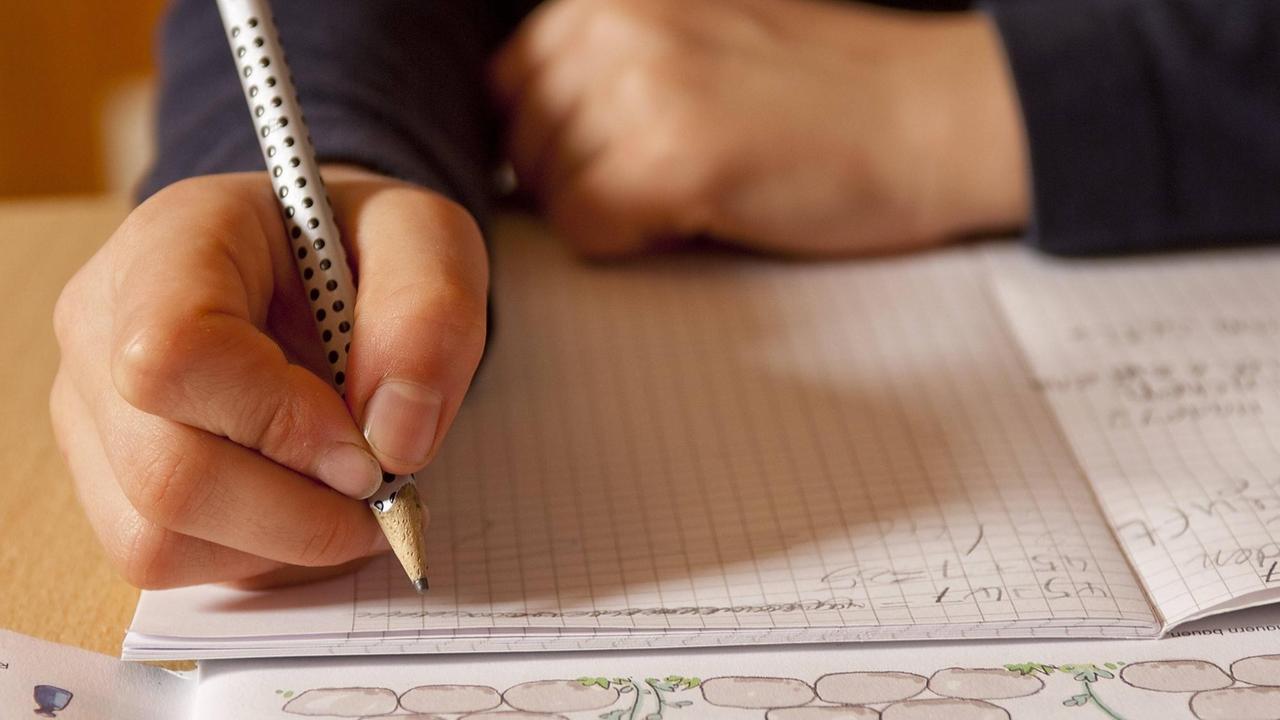 Eine Kind schreibt mit einem Bleistift in einem Hausaufgaben-Heft.