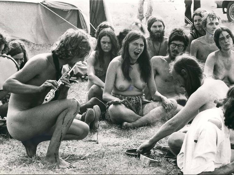 Schwarz-weiß-Aufnahme: eine Gruppe Männer und Frauen, nackt oder halb nackt, sitzen auf einer Wiese bei einem Festival, einer der Männer spielt Querflöte.
