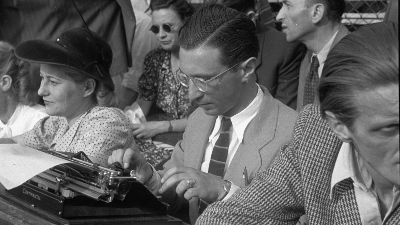 Ein junger Mann mit gescheiteltem Haar, Anzug und Brille sitzt auf der Tribüne eines Stadions und tippt auf einer Schreibmaschine.