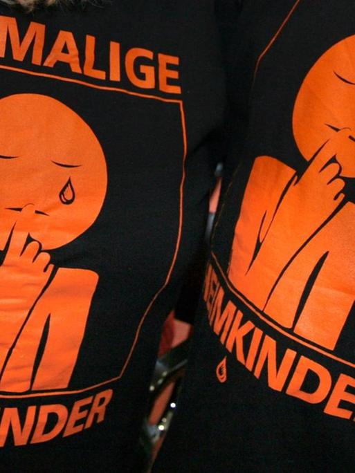 "Ehemalige Heimkinder" ist auf den T-Shirts eines Mannes und einer Frau zu lesen, auf denen auch eine weinende Figur aufgedruckt ist.