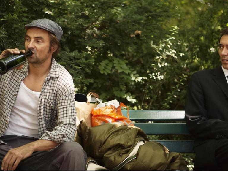 Ein Mann sitzt mit einem Bier und großer Tasche auf der Bank, ein anderer im Anzug schaut ihn spektisch an.