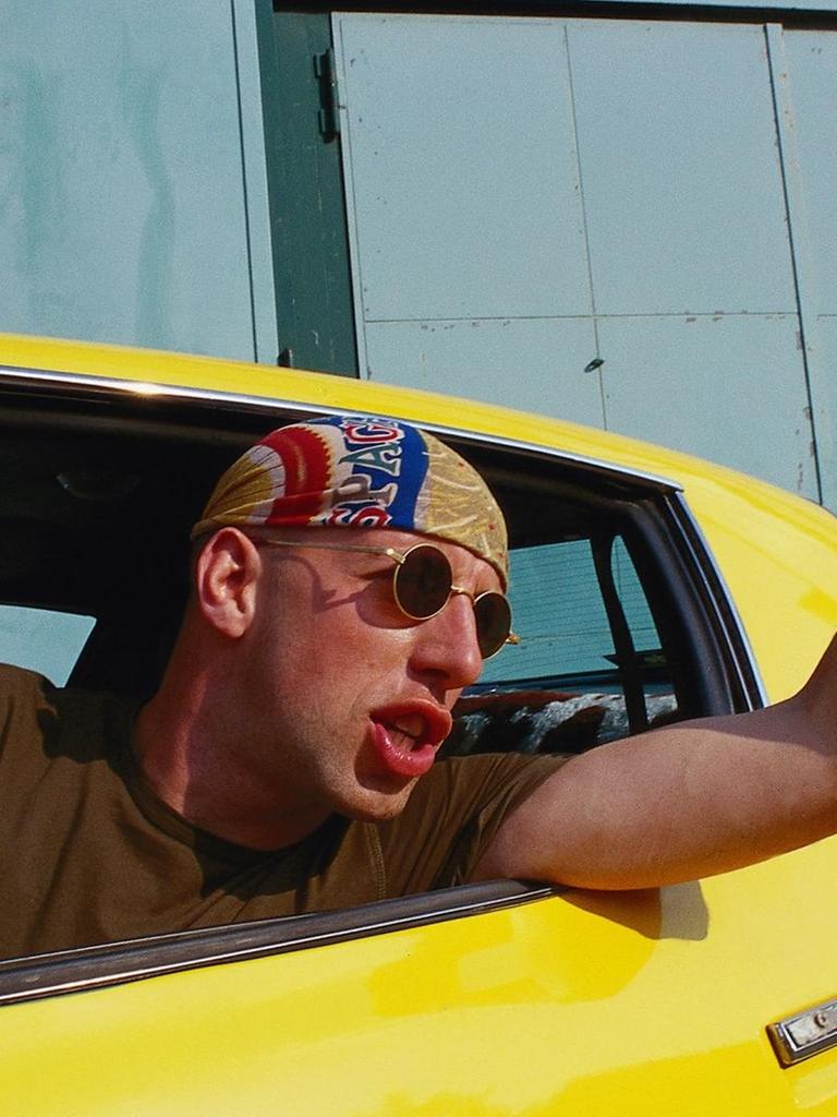 Ein Autofahrer mit Baseball-Kappe reckt seinen Arm aggressiv gestikulierend und schimpfend aus dem Fenster seines gelben Autos.