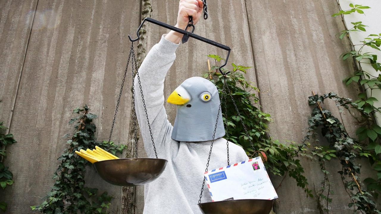 Ein Mensch trägt eine Maske, die einen Taubenkopf darstellt. In der rechten Hand hält die Taube eine Waage, in deren Schalen Briefe und gelbe Hefte liegen.