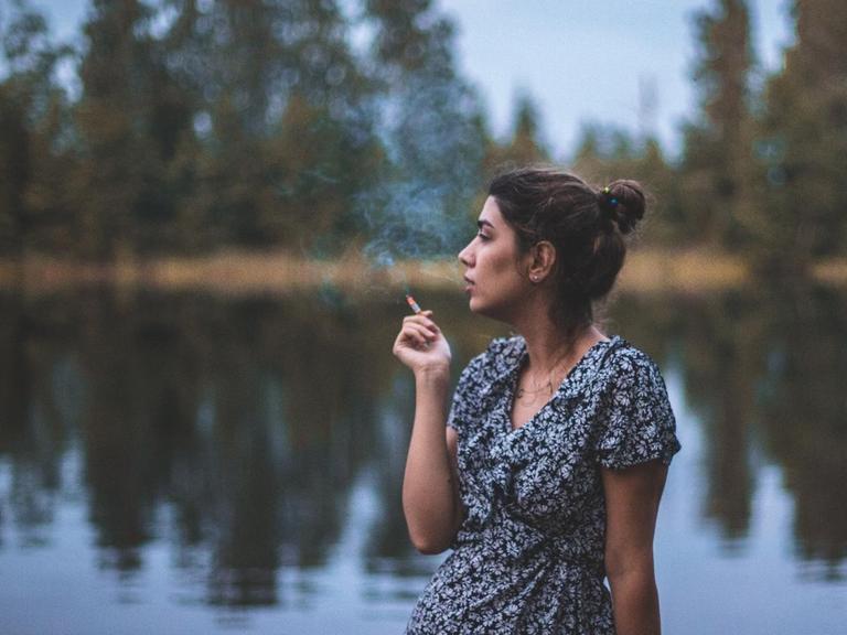Eine junge Frau im Sommer steht an einem See und raucht.
