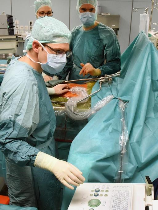 Eine Operation am Transplantationszentrum des Uniklinikums Leipzig. Medizinisches Personal in OP-Kleidung steht am OP-Tisch, eine Person bedient einen Computer.