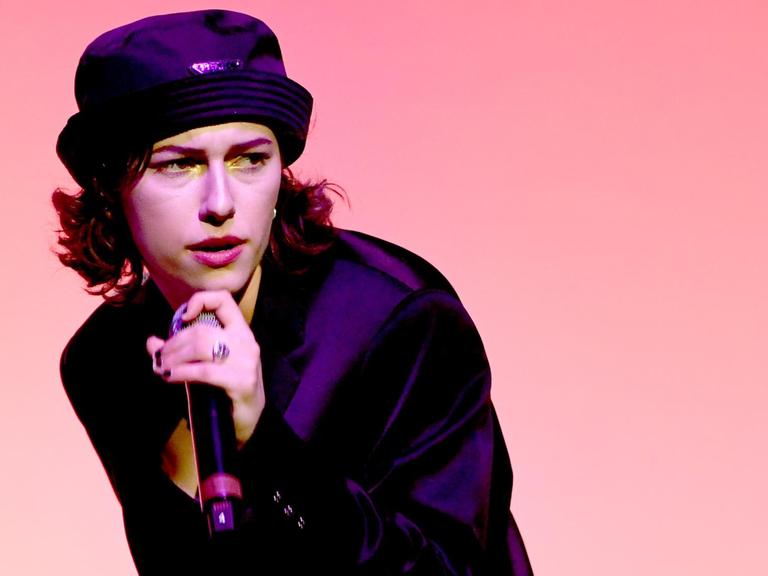 King Princess mit Mikro in der Hand vor einem rosa Hintergrund.