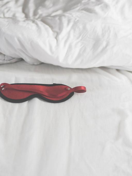 Eine rote, mit schwarzem Rand eingefasste Schlafmaske auf einem weißen Bettlaken. Dahinter ein weiß überzogenes Kopfkissen