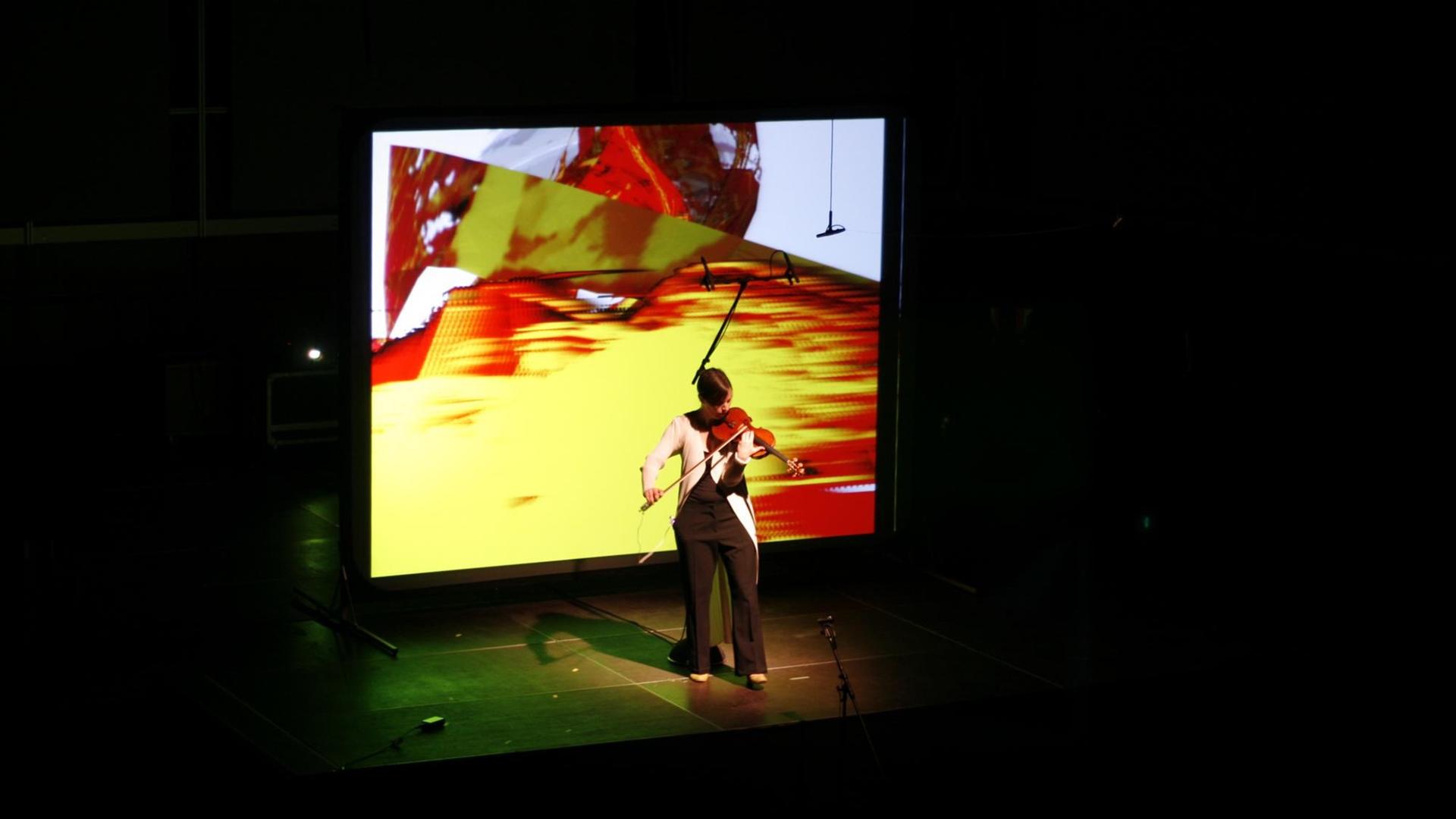 Geigerin Barbara Lüneburg steht mit ihrem Instrument vor einer Videoleinwand Geigerin Barbara Lüneburg steht mit ihrem Instrument vor einer Videoleinwand