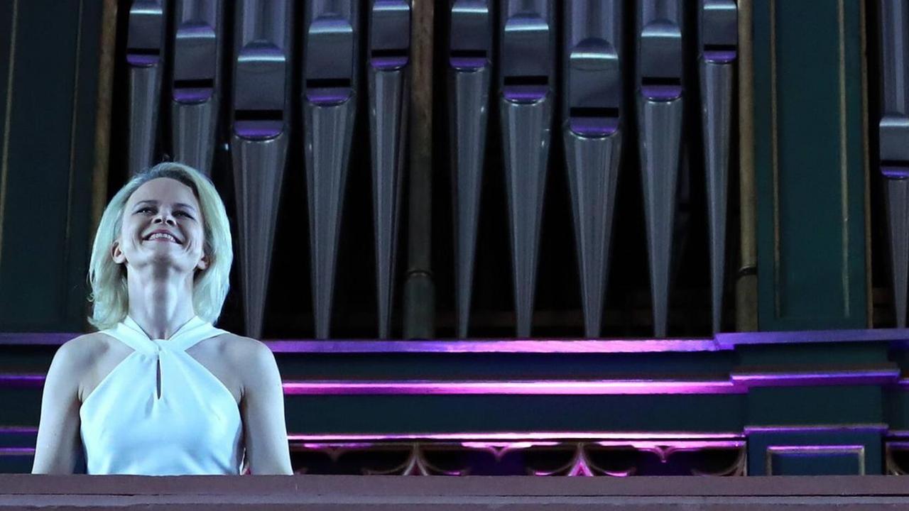 Iveta Apkalna steht in einem weißen Kleid auf einer Empore vor großen Orgelpfeifen. 