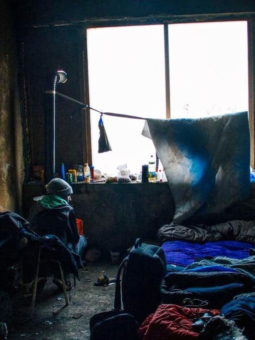 Ein Flüchtling an der bosnisch-kroatischen Grenze sitzt in einem abbruchreifen Haus in einem kalten Zimmer, umgeben von vielen Schlafsäcken.
