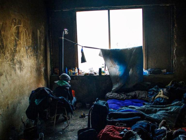 Ein Flüchtling an der bosnisch-kroatischen Grenze sitzt in einem abbruchreifen Haus in einem kalten Zimmer, umgeben von vielen Schlafsäcken.