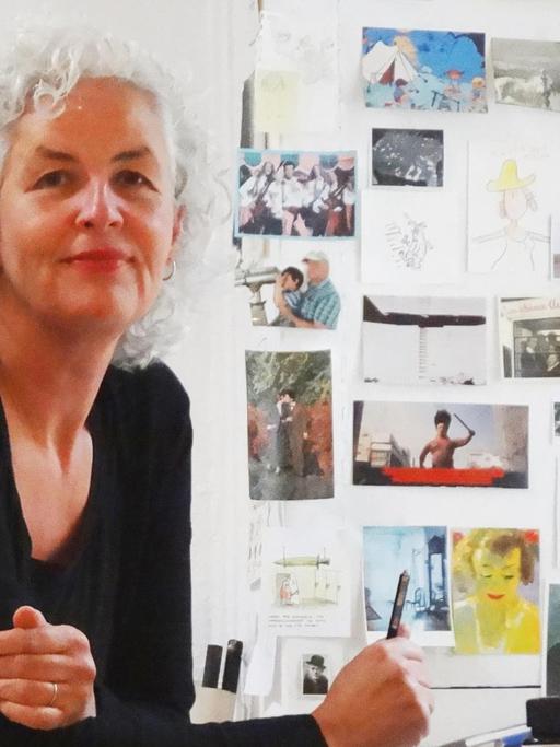 Die Cartoonistin Miriam Wurster sitzt an ihrem Zeichentisch. Um sie herum sind Postkarten, Skizzen und Pinsel zu sehen